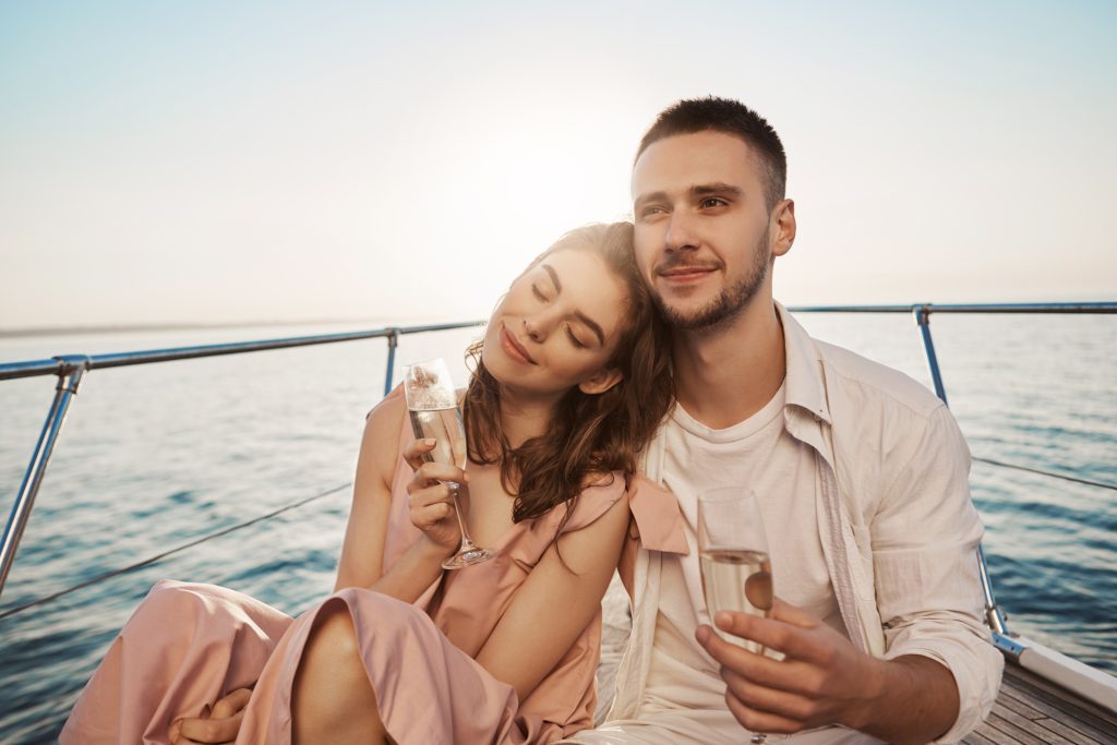 Outdoor portrait of adult happy male and female in love, celebrating their engagement on yacht, holding glass of champagne and hugging. Boyfriend just told about things he likes in her